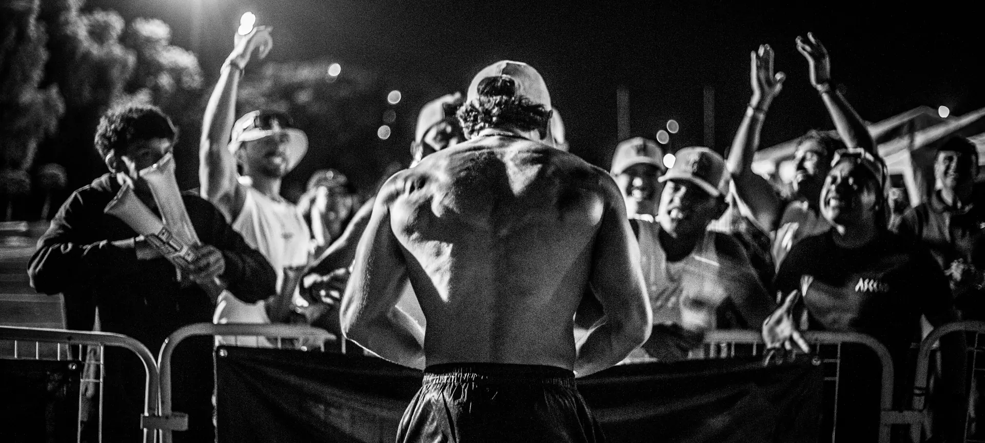 Atleta de espaldas frente al público que lo anima detrás de las vallas, en una competencia al aire libre de noche