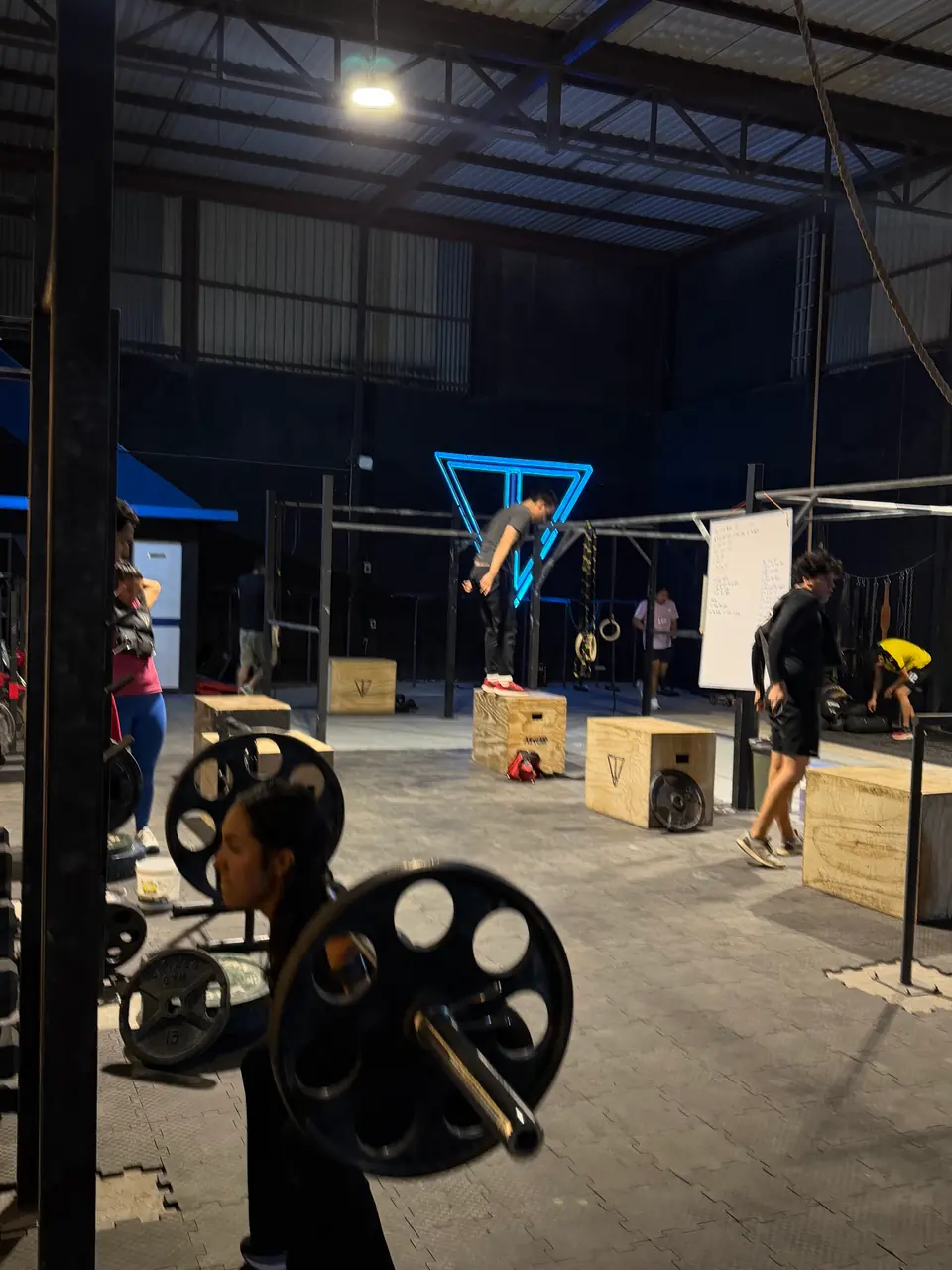 Atleta cargando una barra con discos en primer plano mientras al fondo otros entrenan en la estructura de barras bajo el logo de neón azul