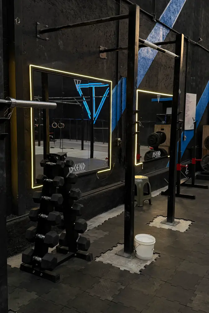 Espejos con marco de luz cálida y rack de mancuernas hexagonales, con el logo de neón azul reflejado