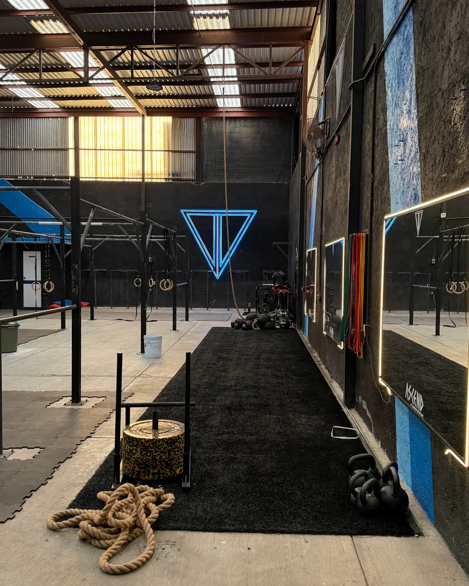 Pasillo de césped negro hacia el logo de neón azul, con kettlebells y un trineo de arrastre cargado con discos